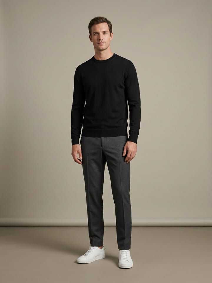 Man in a black merino crewneck and charcoal tapered wool trousers, wearing plain white leather sneakers with no visible branding.