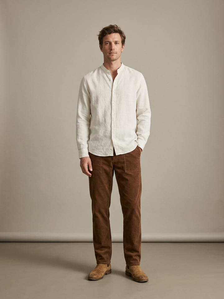 Man in an ecru collarless linen shirt and brown corduroy trousers, wearing tan suede boots.