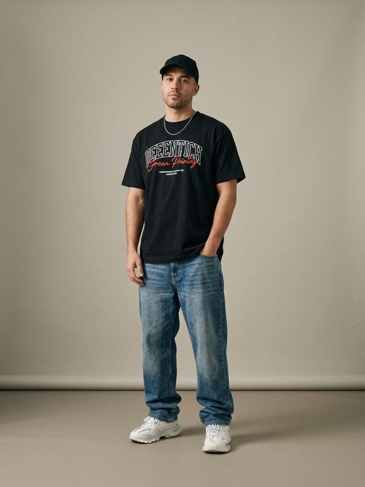 Hombre con vaqueros anchos de lavado claro y una camiseta gráfica negra, llevando zapatillas blancas gruesas y una gorra de béisbol negra.