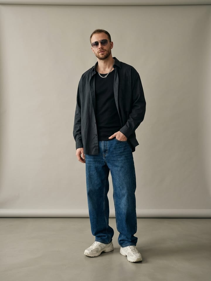 Man in a dark button-down shirt worn open over a black tee with baggy dark jeans, a silver chain necklace and tinted sunglasses.