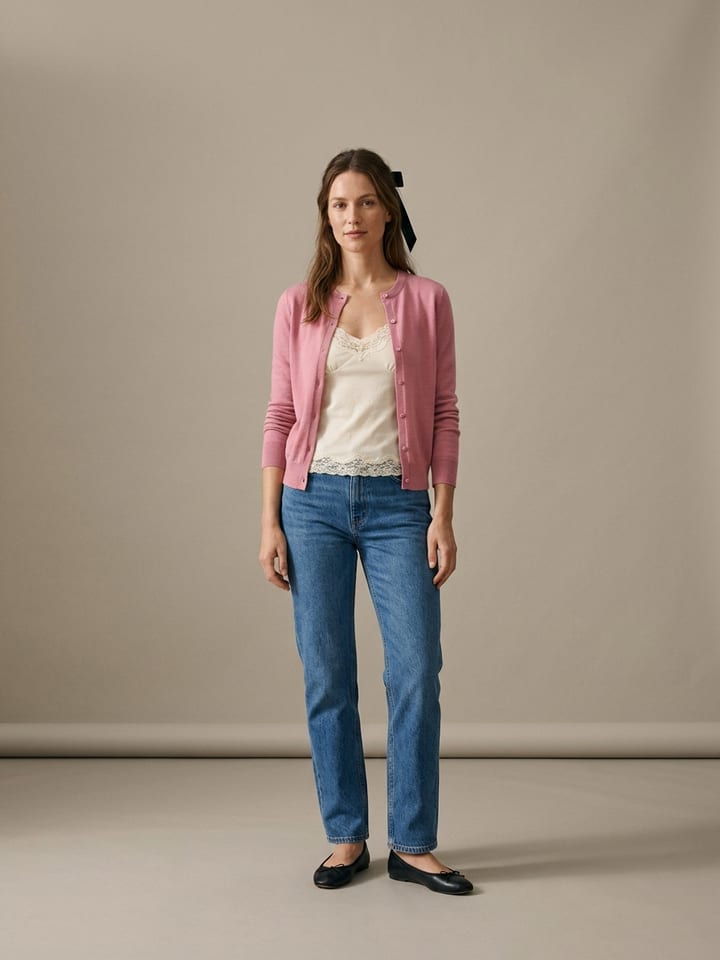 Woman in a cream lace-trimmed cami under a fitted pink cardigan buttoned at the top, with straight blue jeans, black ballet flats and a ribbon tied in her hair.