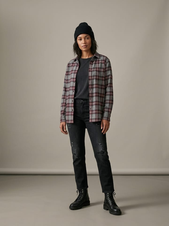 Mulher com uma camisa de flanela xadrez cinzenta e bordeaux aberta sobre uma t-shirt preta desbotada, com jeans pretos retos, botas de atacadores e um gorro preto.