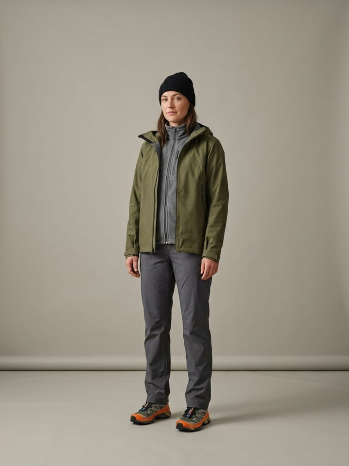 Woman in an olive waterproof shell jacket over a grey zip fleece, with straight charcoal nylon pants, orange-accented trail-running shoes and a black beanie.