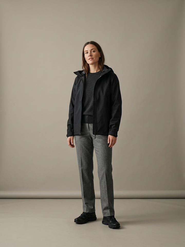 Woman in a black technical shell jacket over a charcoal fine knit, with straight grey wool trousers and clean black trail-style sneakers.