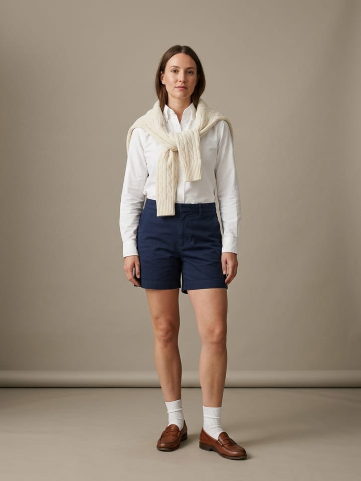 Mujer con una camisa oxford blanca de cuello abotonado y bermudas de sastre azul marino, con un jersey de ochos color crema sobre los hombros y mocasines penny marrones.