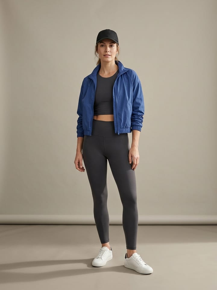 Woman wearing a matching charcoal legging and crop top set under a cropped cobalt blue windbreaker, with low-profile white sneakers and a black cap.