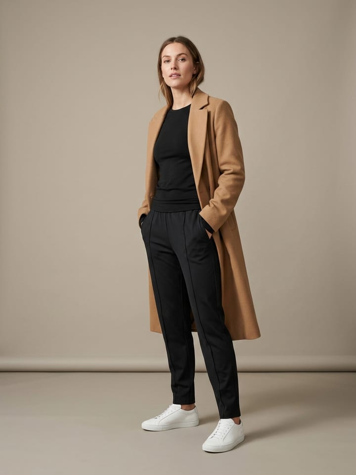 Woman in sleek black track pants and a fitted black long-sleeve top under an open longline camel wool coat, wearing clean white sneakers.