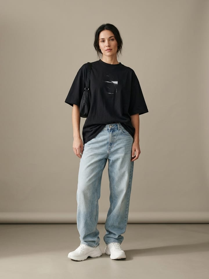 Mujer con vaqueros anchos de lavado claro y una camiseta gráfica negra oversize remetida por delante, llevando zapatillas blancas gruesas y un pequeño bolso de hombro.