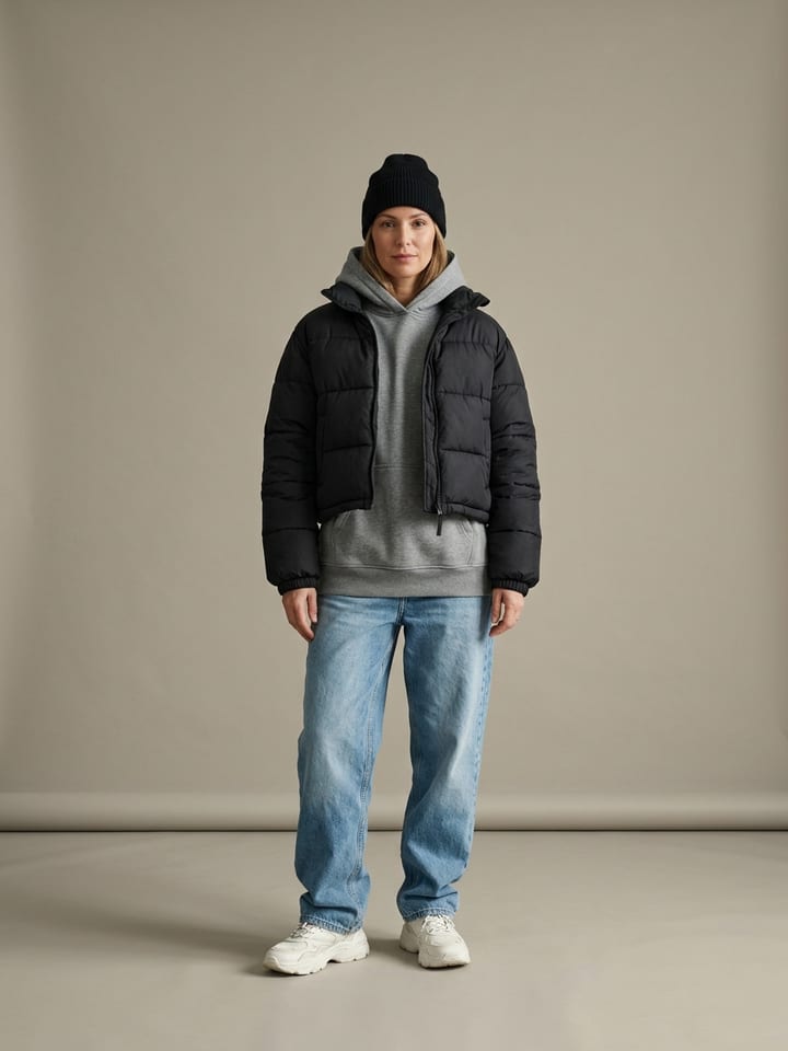 Mujer con una chaqueta plumífero negra corta sobre una sudadera con capucha gris, con vaqueros anchos, zapatillas gruesas y un gorro de lana negro.